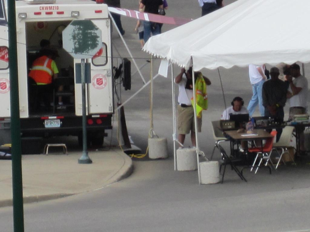 Ein Rettungswagen steht auf der linken Seite einer Straße, während auf der rechten Seite ein Zelt viele Menschen schützt, bei denen ein Tisch und ein Stuhl in der Nähe stehen.