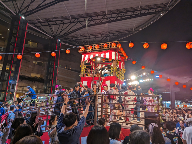 Menschenmenge auf einer beleuchteten Bühne mit Laternen, einige halten Kameras, umgeben von einem Geländer, mit Bäumen und einem klaren blauen Himmel im Hintergrund, möglicherweise während eines japanischen Festivals.