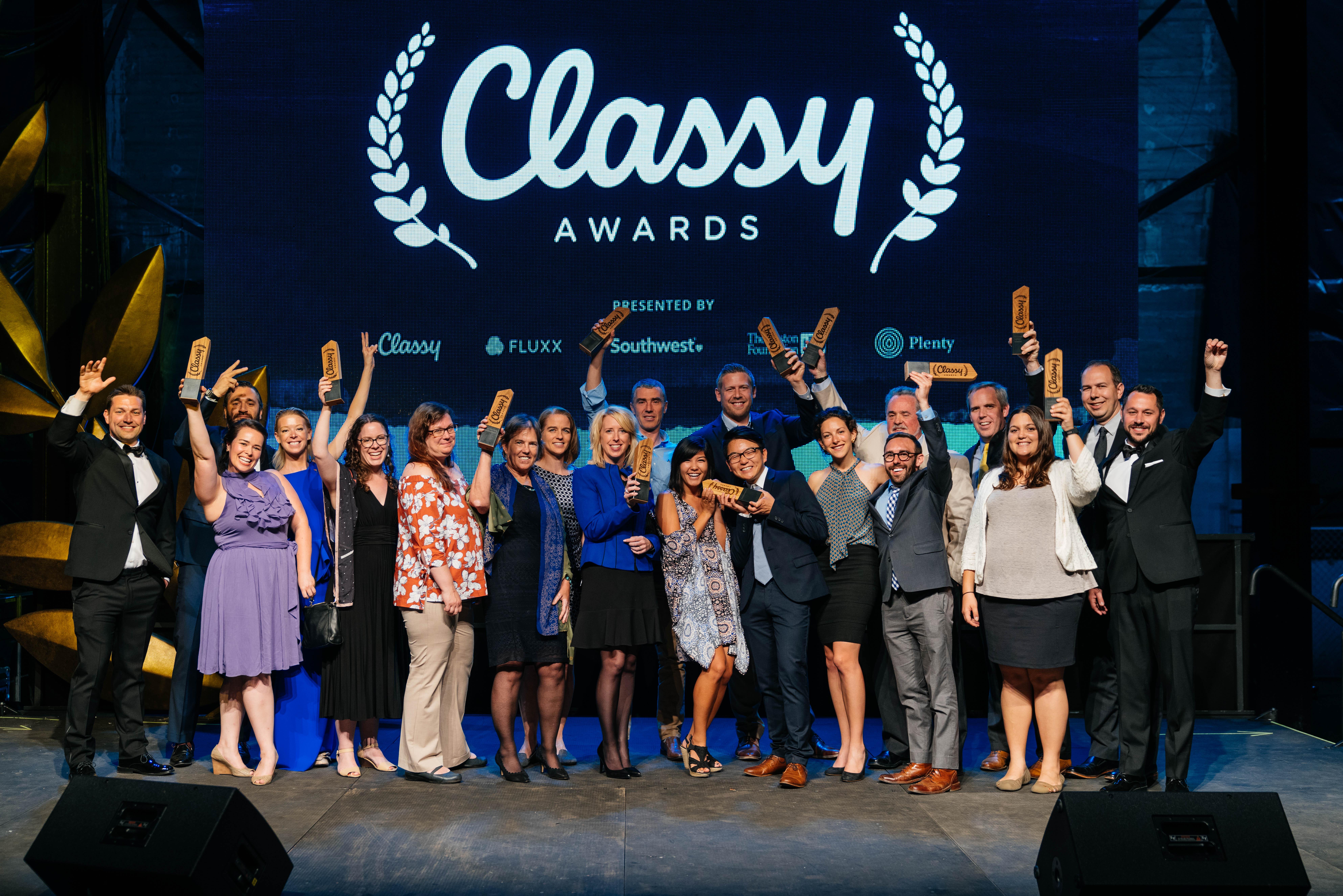 Eine Gruppe von Menschen auf einer Bühne, die lächeln und Auszeichnungen halten, mit Lautsprechern unten und einem Bildschirm im Hintergrund, auf dem 'Classy Awards 2019' angezeigt wird.