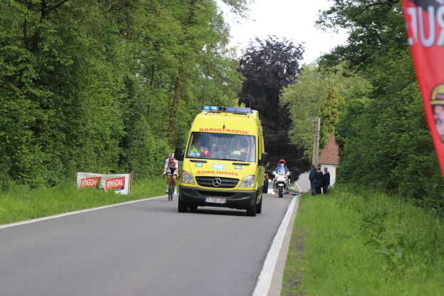 Ambulanz fährt auf Straße mit Fahrradfahrern nebenher, Gras und Bäume auf beiden Seiten, Häuser und Masten im Hintergrund unter einem klaren blauen Himmel.