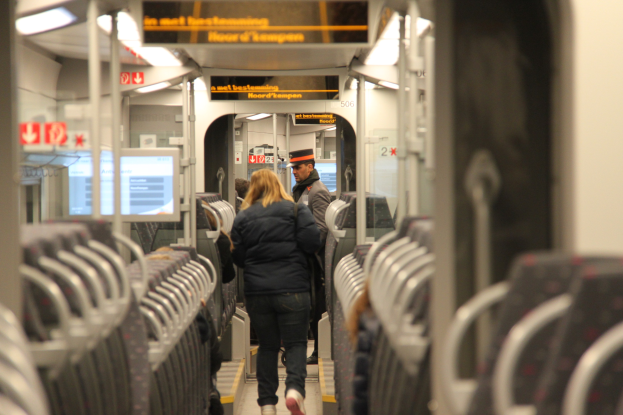 Eine Gruppe von Menschen, die in einem U-Bahn-Zug gehen, mit einem Mann und einer Frau im Vordergrund; das Innere des Zugs enthält Sitzplätze, Stangen, Lampen, Texttafeln, Bildschirme und andere Gegenstände.