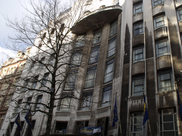 Gebäude mit Fenstern, einer Fahne und Flaggen an Stangen unten und einem Baum mit Zweigen.