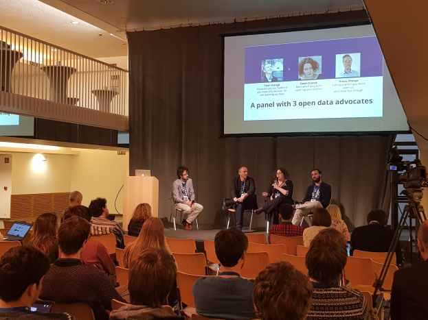 Eine Gruppe von Menschen, die auf Stühlen vor einem Projektionsschirm sitzen, mit einem Laptop auf einem zentralen Tisch, einer Kamera auf einem Ständer rechts daneben und Geländern, Lampen und einer Wand im Hintergrund während einer Podiumsdiskussion über Open-Data-Aktivisten.
