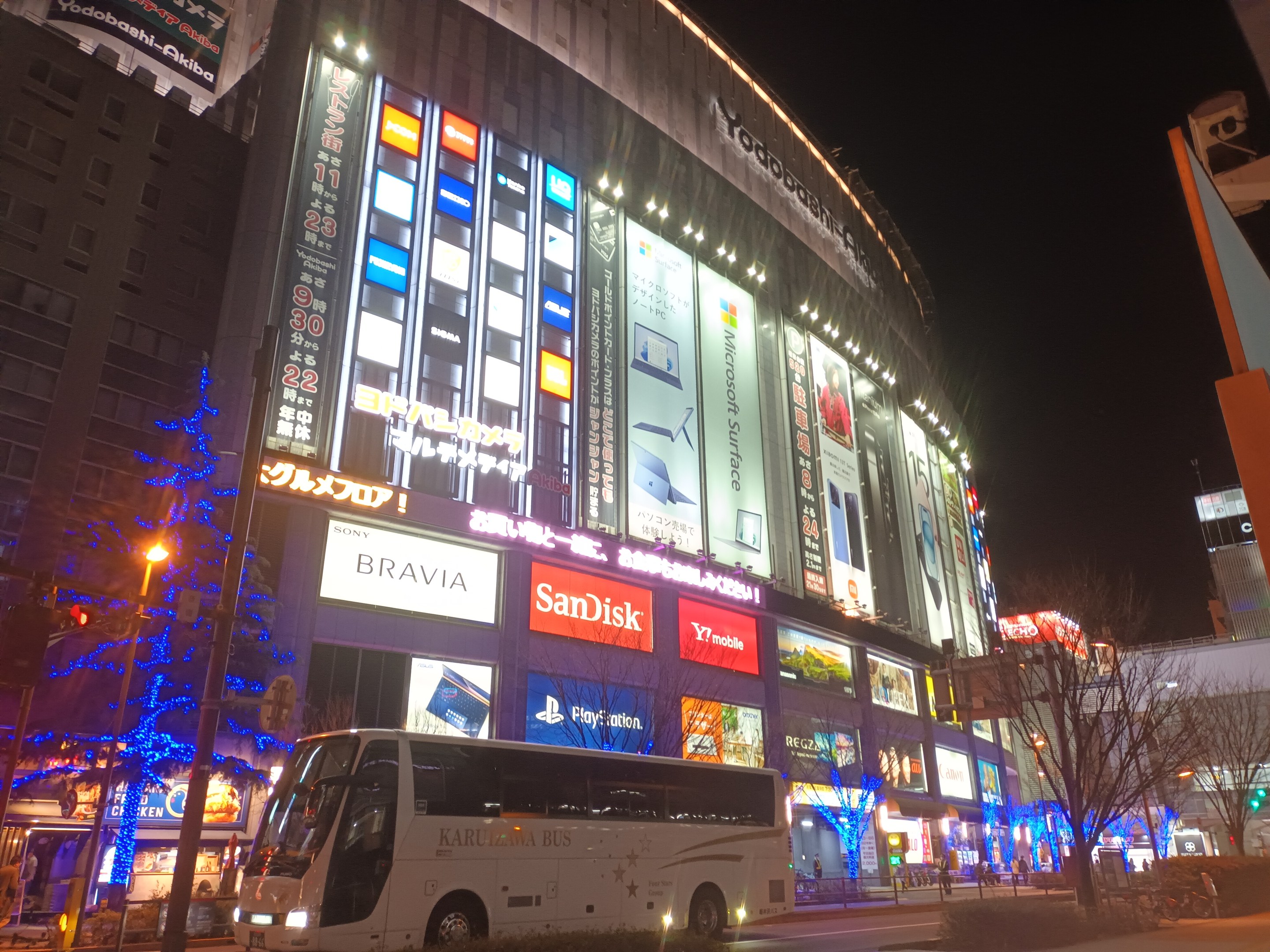 Ein Bus fährt nachts auf einer Stadtstraße, umgeben von hohen Gebäuden mit Weihnachtsbeleuchtung, Straßenlaternen, Bäumen und verschiedenen städtischen Elementen unter einem sichtbaren Himmel.