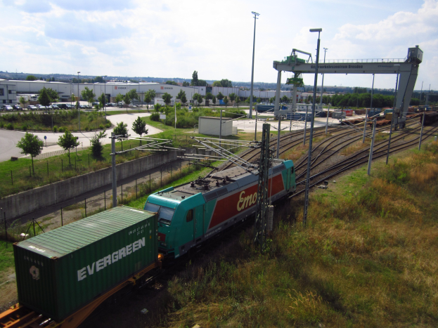 Güterzug, der auf Schienen neben einem grünen Feld mit Strommasten, Kabeln, Bäumen, Gebäuden, Fahrzeugen und einem bewölkten Himmel im Hintergrund fährt.