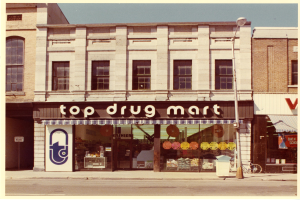 Eckladen namens "Top Drug Mart" mit Fenstern, einem Schild, einem Straßenschild, einem parkenden Fahrrad, einem Müllcontainer und einem bewölkten Himmel.