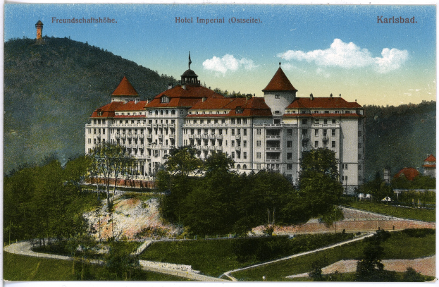Eine alte Postkarte des Hotel Imperial in Karlsbad, Deutschland, zeigt ein großes Gebäude mit Fenstern, umgeben von Bäumen, Pflanzen, Gras, einem Pfad, einer Baumgruppe, Hügeln und einem bewölkten Himmel, mit Text oben.