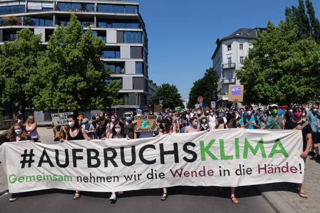 Eine Gruppe von Menschen mit Masken hält ein Banner mit der Aufschrift 'Aufbruchsklima' vor einem Gebäude, umgeben von Bäumen unter einem klaren Himmel.