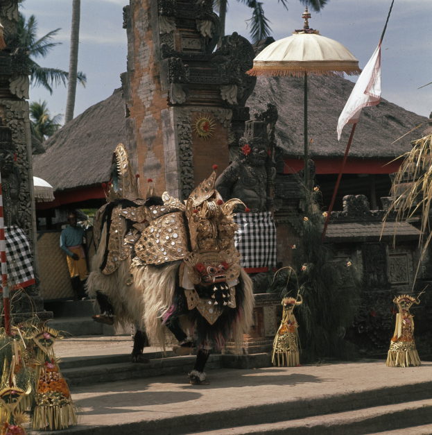 Eine Gruppe von Menschen in traditionellen balinesischen Kostümen tanzt vor einem Tempel mit Treppen, einem Fahnenmast, Hütten und Bäumen im Hintergrund unter einem bewölkten Himmel.