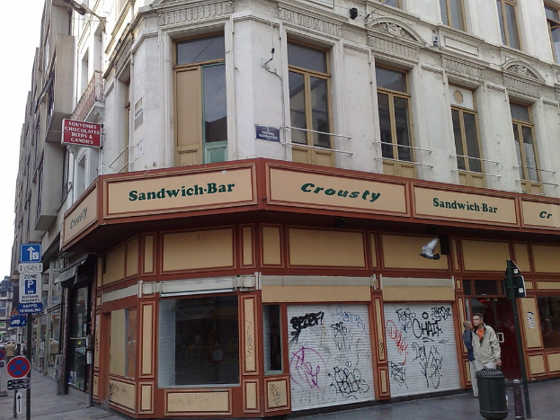 Ein Gebäude mit Wänden, Fenstern, Glastüren, Geländern, Namensschildern und Geschäften, das eine Stange auf der rechten Seite, einen Müllcontainer und Stangen mit Schildern auf der linken Seite zeigt.