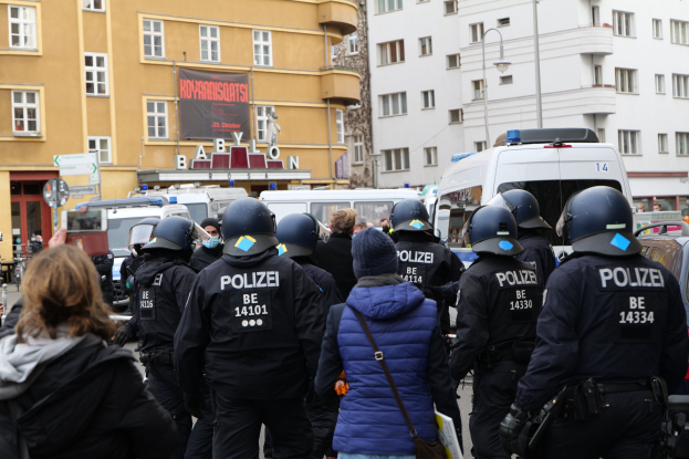 Polizeibeamte in Uniform vor einer Menge mit Helmen und Jacken bei einer Demonstration in Berlin, Deutschland, mit Fahrzeugen, Gebäuden, Laternenmasten und einem Transparent im Hintergrund.