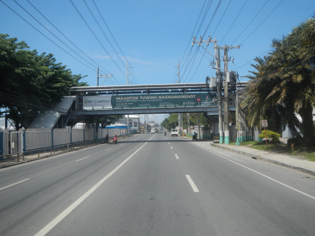 Arterielle Straße mit Fahrzeugen, Strommasten und -leitungen, eine Brücke mit Namensschild, Bäume auf beiden Seiten, ein Zaun, Gebäude und ein bewölkter Himmel.