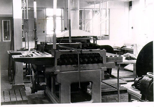 Ein Schwarz-Weiß-Foto eines Druckers in einer Fabrik, umgeben von Maschinen, Polen, Fenstern und einer Wand im Hintergrund.