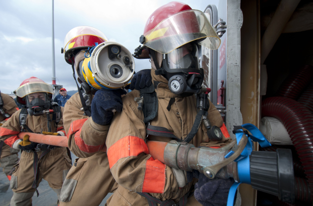 Feuerwehrleute in Schutzausrüstung, einer hält einen Schlauch, mit einer Stange, Rohren und bewölktem Himmel im Hintergrund.