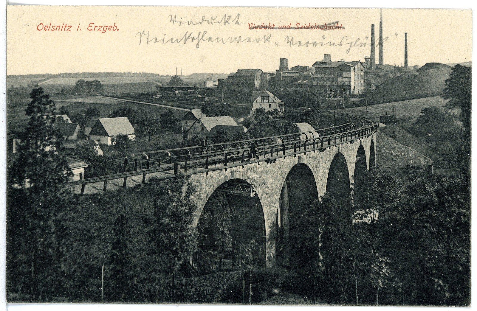 Schwarzes und weißes Foto einer Brücke mit mehreren Bögen und Pfeilern, einem Zug, der darüber fährt, und mehreren Menschen, die auf der Brücke stehen; im Hintergrund sind Bäume, Gebäude, Strommasten und Himmel zu sehen; oben auf dem Bild ist Text zu erkennen.