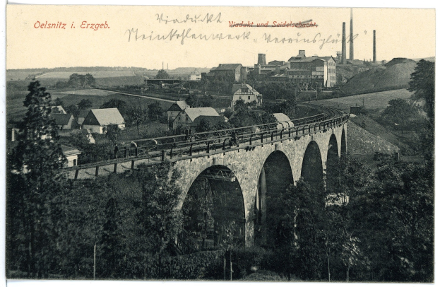 Schwarzes und weißes Foto einer Brücke mit mehreren Bögen und Pfeilern, einem Zug, der darüber fährt, und mehreren Menschen, die auf der Brücke stehen; im Hintergrund sind Bäume, Gebäude, Strommasten und Himmel zu sehen; oben auf dem Bild ist Text zu erkennen.