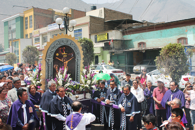 Menschen auf einer Straße außerhalb der Stadt, mit einem Kruzifix und Blumensträußen im Vordergrund, Autos im Hintergrund und Gebäuden, Bäumen und Straßenlaternen weiter hinten.