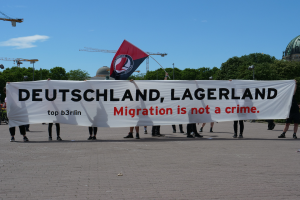 Eine Gruppe von Menschen hält eine Fahne mit der Aufschrift "Deutschland, Lagerland Migration ist kein Verbrechen" und eine Flagge, mit Straßenlaternen, Bäumen, Kränen, einem Gebäude und einem bewölkten Himmel im Hintergrund.