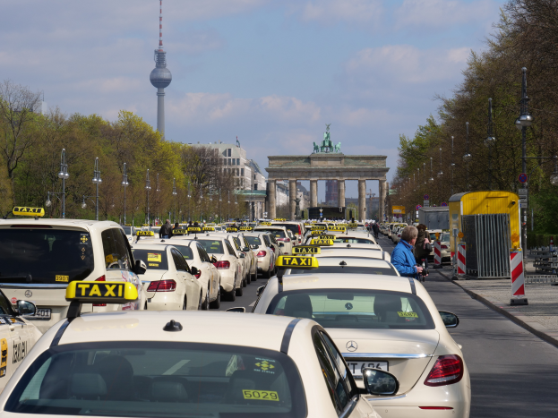 Eine belebte Straße in Berlin, Deutschland, mit vielen parkenden Taxis, Fußgängern auf dem Gehweg, Laternen, Bäumen, Gebäuden und einem fernen Bogen mit Statuen und einem Turm unter einem bewölkten Himmel.
