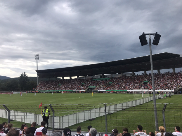 Große Zuschauermenge bei einem Fußballspiel in einem Stadion mit bewölktem Himmel und beleuchtetem Feld, umgeben von einem Zaun, Bäumen und Hügeln.