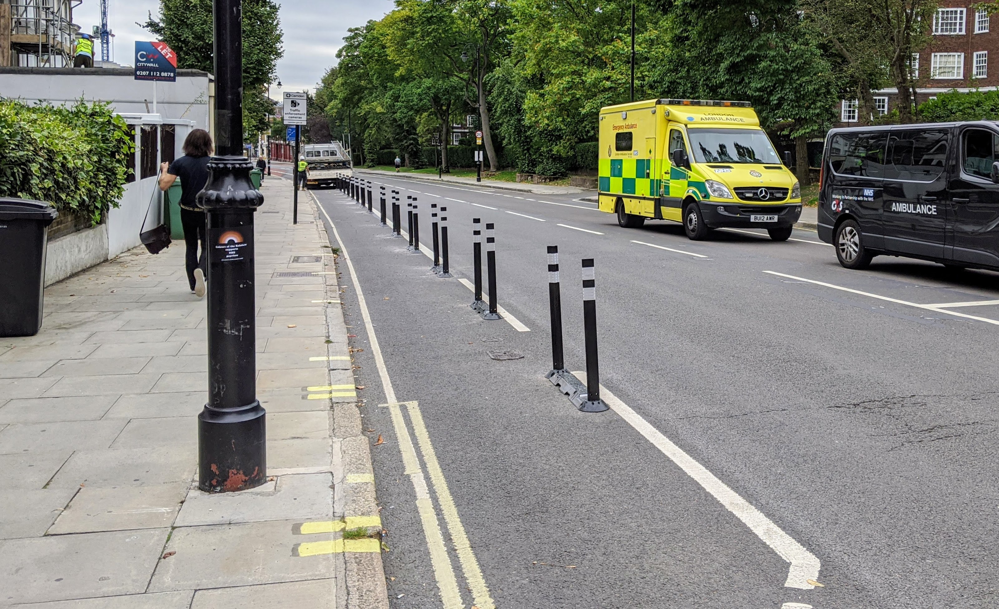 Ambulanz auf einer Straße neben einem Gehweg mit Fahrzeugen, einem Fußgänger, einem Müllcontainer, Versorgungsmasten, Schildern, Bäumen, Gebäuden und einem bewölkten Himmel.
