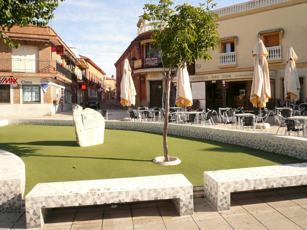 Minigolf-Platz in einem Stadtplatz mit umgebenen Sitzgelegenheiten, Schirmen, Bäumen und Gebäuden unter einem bewölkten Himmel.