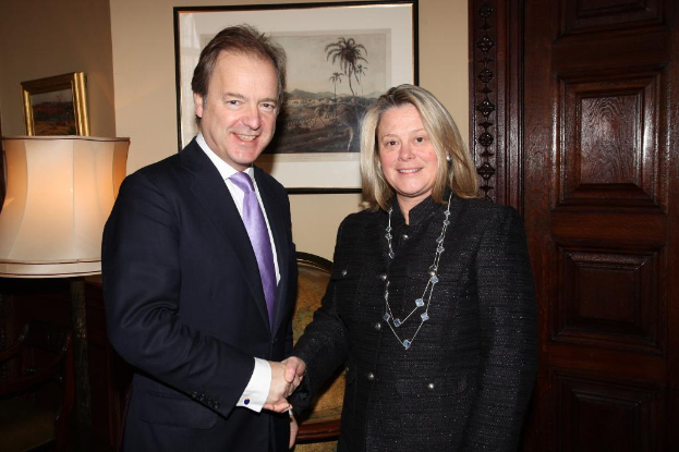 Eine Frau und ein Mann in einem Anzug lächeln, schütteln sich die Hände und sehen sich an, mit einer Wand, Fotorahmen, einer Lampe, einer Tür und einigen Gegenständen im Hintergrund.