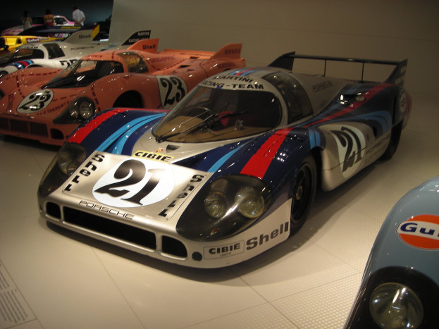Ein Porsche 917K Le Mans-Rennwagen wird in einem Museum ausgestellt, umgeben von anderen Sportwagen und Menschen, mit einer Wand im Hintergrund und einem Papier mit Text auf der linken Seite.
