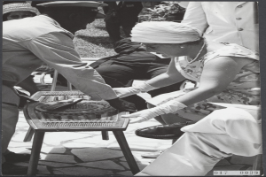 Ein Schwarz-Weiß-Foto eines Mannes und einer Frau, die Backgammon an einem Tisch spielen, umgeben von Menschen, Pflanzen und Gras.