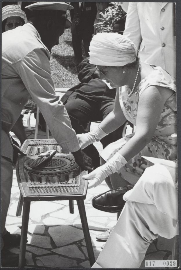 Ein Schwarz-Weiß-Foto eines Mannes und einer Frau, die Backgammon an einem Tisch spielen, umgeben von Menschen, Pflanzen und Gras.
