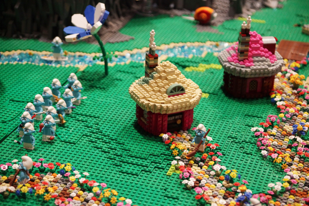 Ein LEGO-Modell einer Kleinstadt mit bunten Blumen, Häusern, Pflanzen und Spielzeugen vor einem leicht unscharfen, träumerischen Hintergrund.