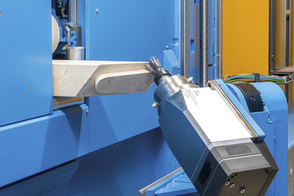 Eine CNC-Fräsmaschine schneidet ein Holzstück in einer Fabrikumgebung, umgeben von Kabeln und anderer Ausrüstung, mit einer Wand im Hintergrund.