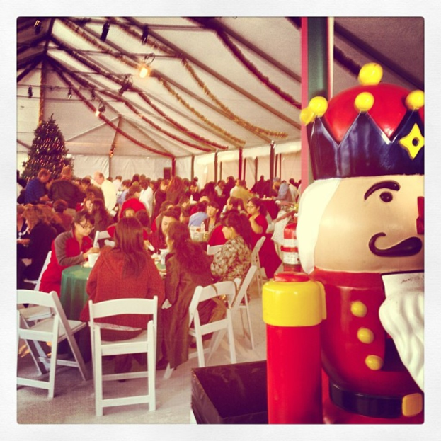 Eine Gruppe von Menschen feiert Weihnachten in einer Hütte, mit einem geschmückten Baum und weißen Stühlen, einer roten Spielzeug-Sicherheitswache mit schwarzer Krone.