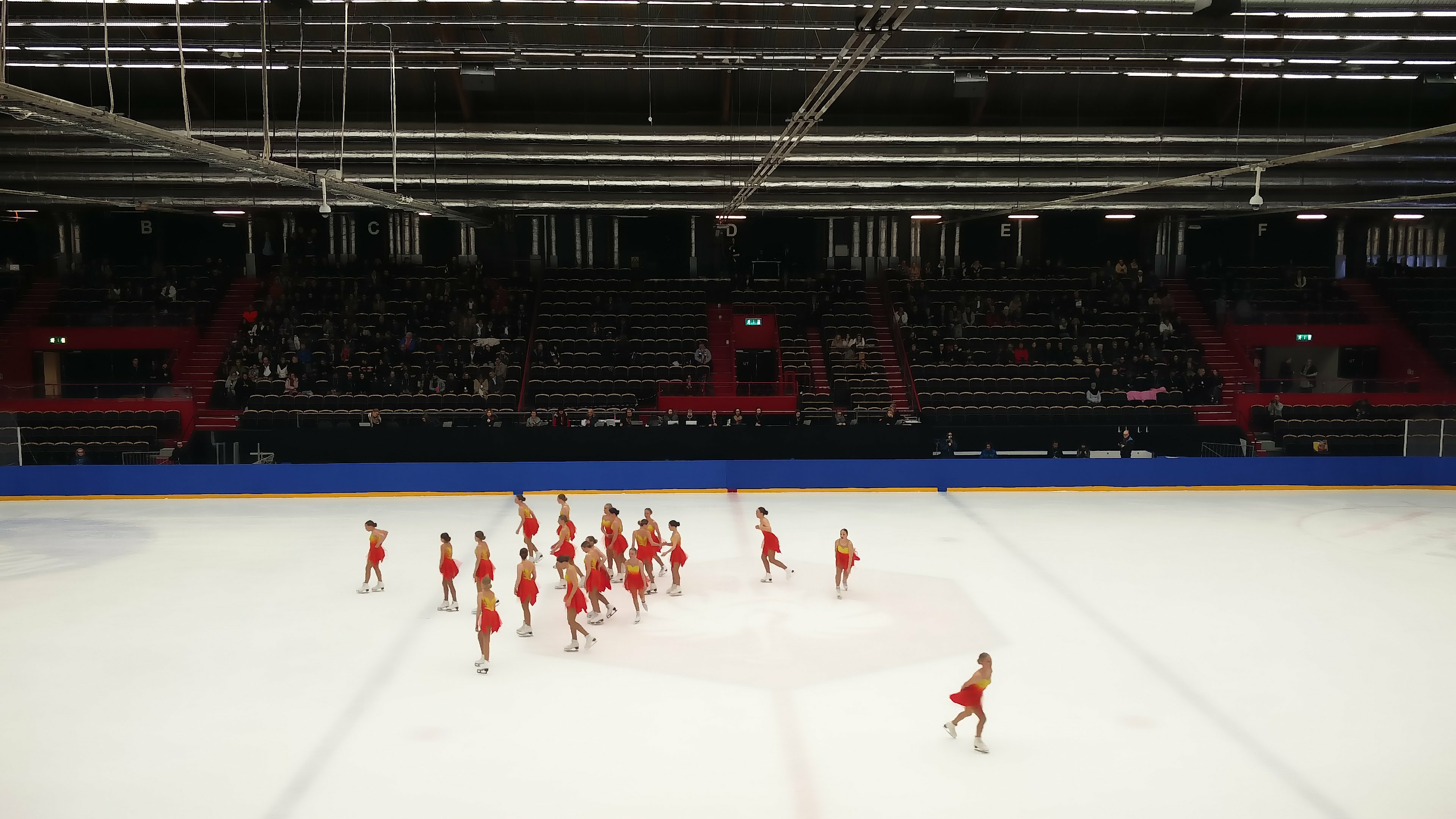 Eine Gruppe von Menschen, die auf einer Eisbahn Schlittschuh laufen, umgeben von einem Publikum, das auf Stühlen sitzt, mit Scheinwerfern, die die Szene beleuchten.