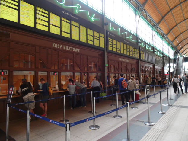 Menschen, die in einer Warteschlange an einem Bahnhofsschalter mit Leuchten, Informationsanzeigen und bannergeschmückten Pfählen stehen.