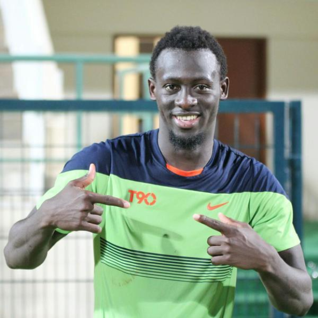 Ein Mann in einem grün-blauen Hemd, identifiziert als Emmanuel Adebayor, zeigt auf die Kamera und lächelt, mit einem unscharfen Geländer, einer Wand und einer Tür im Hintergrund.