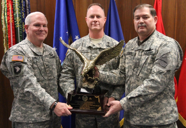 Drei Männer in Uniformen stehen mit Auszeichnungen, Flaggen im Hintergrund und buntem Text in der linken oberen Ecke.