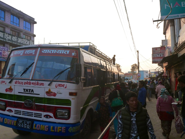 Eine Gruppe von Menschen, die in einem städtischen Gebiet stehen und gehen, mit einem Bus, Texttafeln, Gebäuden, einem Baum und einem Strommast mit Drähten im Hintergrund.