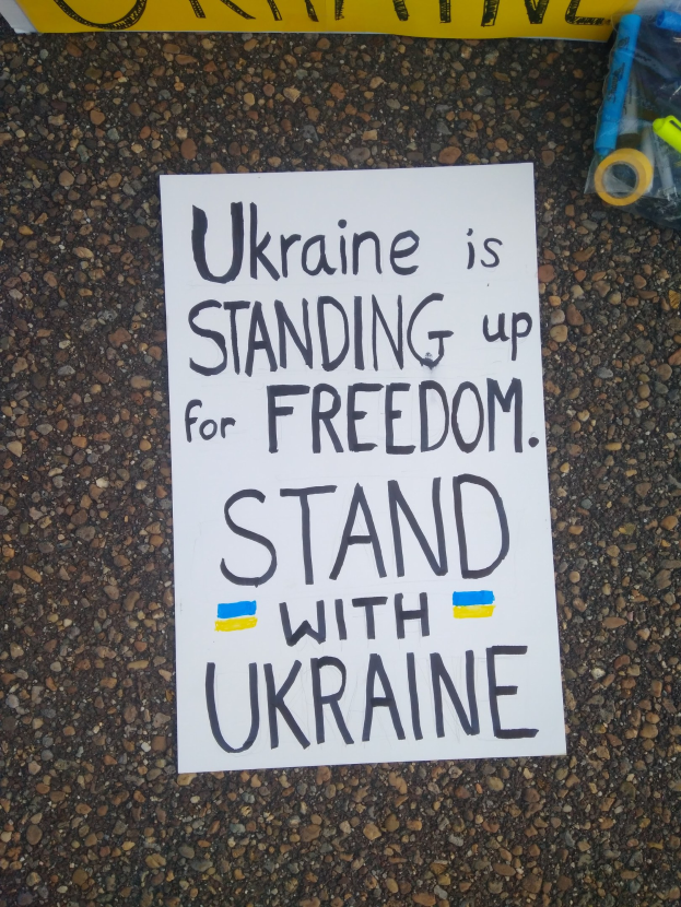 Ein weißer Zettel mit der Aufschrift "Ukraine steht für Freiheit auf, stellt dich mit Ukraine" liegt vor einer gelben Fahne auf dem Boden. Rechts davon befindet sich eine Plastikabdeckung mit verschiedenen Gegenständen.