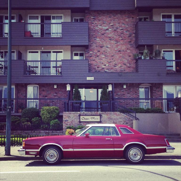 Ein rotes Auto befindet sich unten im Bild, mit einem Gebäude in der Mitte, Pflanzen davor und einem Pfahl auf der linken Seite.
