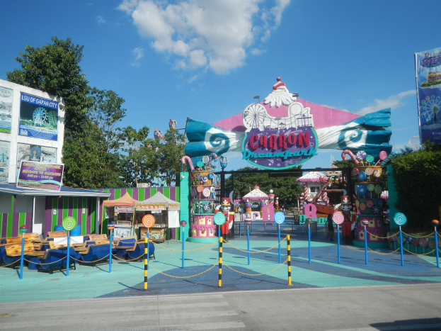 Eine Karussellfahrt am Eingang eines Parks mit einer Straße, einem Zaun, Buden, Banner, Bäumen und einem bewölkten Himmel im Hintergrund.