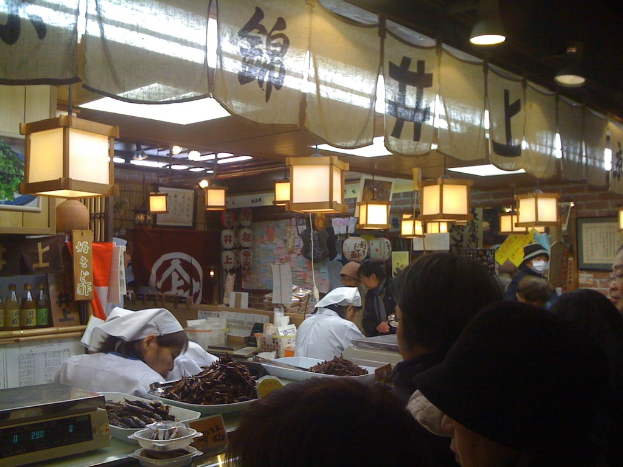 Menschen bei der Arbeit in einem Restaurant mit Kunden an der Theke, verschiedene Gegenstände und Dekorationen im Hintergrund.