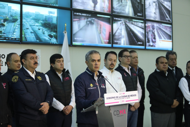 Gruppe von Männern, die vor einem Podium mit einem Mikrofon und einer Tafel mit der Aufschrift 'Mexikanische Zoll- und Grenzschutzbehörde' stehen, mit Fernsehgeräten und einer Flagge an der Wand dahinter.