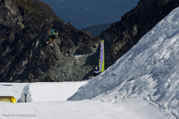Ein Mann in Skier springt in der Luft mit schneebedeckten Bergen und einer Flagge mit Text im Hintergrund und einem anderen Mann vor der Flagge, mit Text in der unteren linken Ecke.
