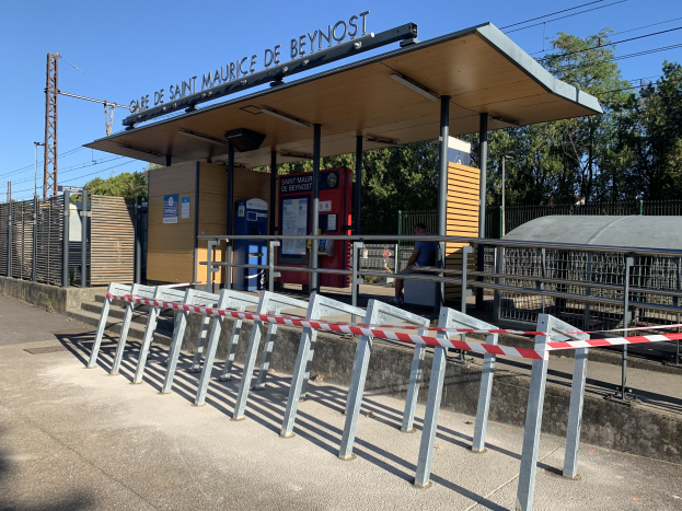 Ein Bahnhofsgebäude mit einem Metallzaun und einem rot-weißen Streifenbarrieren, eine Person auf einer Bank unter einem Vordach sitzend, Bäume, Strommasten mit Drähten und einen klaren blauen Himmel im Hintergrund.