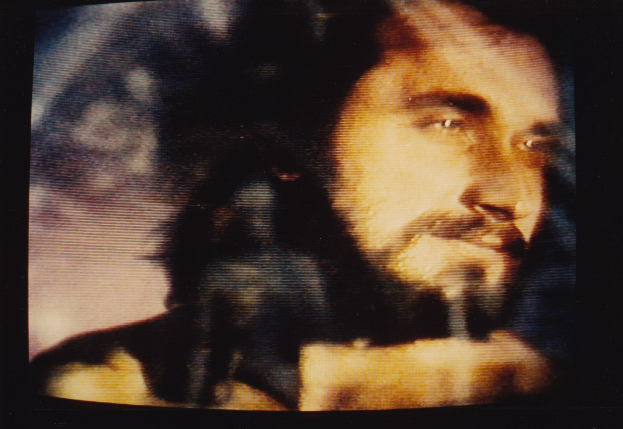 Ein Mann mit Bart und Schnurrbart, langen dunklen Haaren und einem ernsten Gesichtsausdruck, der direkt in die Kamera schaut, mit einem dunklen Hintergrund dahinter.