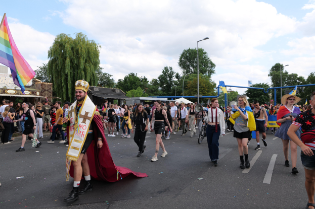 Eine Gruppe von Menschen, die bei der Gay Pride Parade 2018 marschieren und eine Regenbogenflagge sowie Musikinstrumente tragen, mit einigen, die Mützen tragen, an Laternen, Bäumen und Schuppen vorbei unter einem bewölkten Himmel.