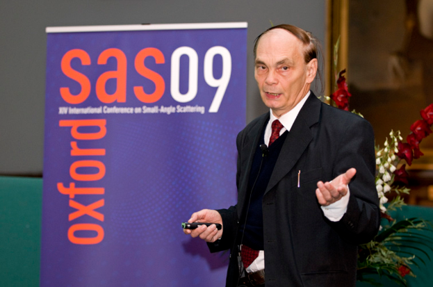 Ein älterer Mann in einem Anzug hält ein Objekt, während er spricht, mit einem Banner, einer Wand, einem Foto Rahmen und Blumen im Hintergrund.