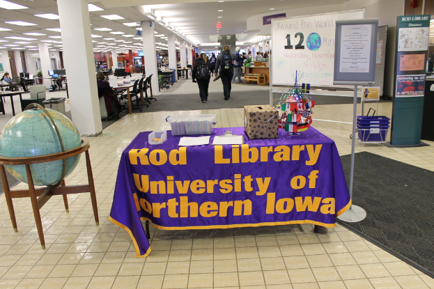 Eine Ausstellung mit einem Tisch, auf dem verschiedene Landesflaggen, eine Box, Papiere und ein Banner liegen, links daneben eine Weltkugel, rechts ein Anschlagbrett mit einigen Menschen in der Nähe und mehreren Tischen und Stühlen links.