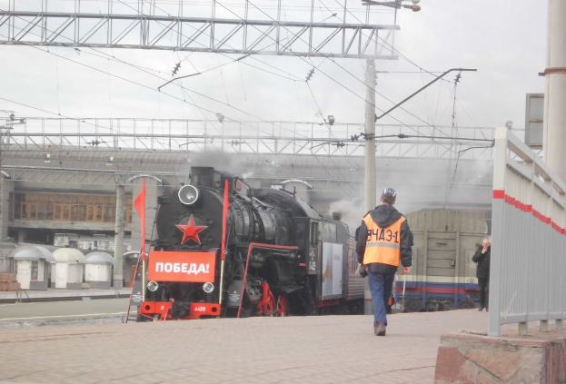 Ein Mann mit Helm und einer Handtasche geht auf einem Bahngleis neben einem Zug, mit einer Wand, einer Sauele, Strommasten mit Drahten, einem Gebaeude und dem Himmel im Hintergrund.